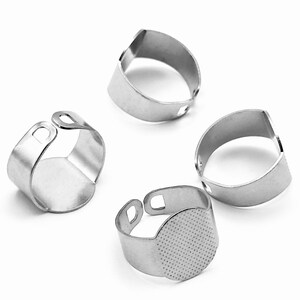 May include: Four silver-colored metal ring blanks with adjustable bands. Each ring features a flat, circular pad at the front for attaching decorations. The bands have an open design with small rectangular cutouts at the ends for adjustment.