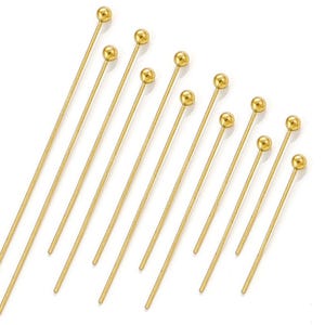 Può includere: Una collezione di spilli dorati con una pallina in cima. Gli spilli sono disposti diagonalmente su uno sfondo bianco. Gli spilli sono di diverse lunghezze, con un gambo sottile e dritto.