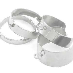 May include: A collection of silver-colored metal bracelets. Each bracelet is a thick band with a smooth, reflective surface. Some bracelets have small, circular attachments. The bracelets are stacked and arranged on a white background.