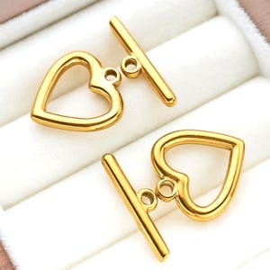 May include: Two gold-coloured heart-shaped toggle clasps. Each clasp features a heart-shaped loop and a straight bar with a small loop. The clasps are displayed on a white, textured surface.