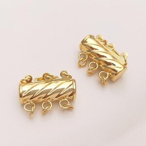 May include: Two gold-toned cylindrical jewellery clasps featuring a diagonal ribbed texture. Each clasp is designed with three small circular jump rings attached to the bottom, allowing for the connection of multiple strands of jewellery or necklaces.