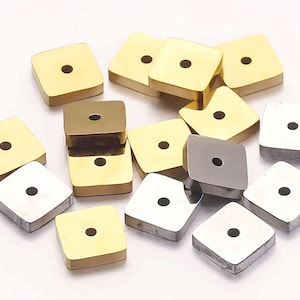 Könnte beinhalten: Eine Sammlung quadratischer Metallperlen in Gold- und Silbertönen. Jede Perle hat ein kleines, mittiges Loch. Die Perlen sind auf einer weißen Oberfläche angeordnet, was ihre polierte Oberfläche und geometrische Form hervorhebt. Ideal für die Schmuckherstellung.