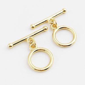 Könnte beinhalten: Zwei goldfarbene Toggle-Verschlüsse mit runden Ringen. Jeder Verschluss hat eine Stange, die durch den Ring gleitet.
