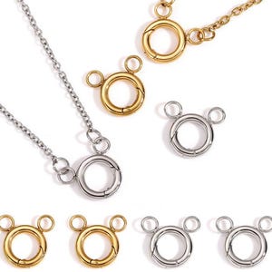Pode incluir: Uma coleção de fechos de joias e colares de corrente nas cores ouro e prata. Os fechos são circulares com dois pequenos laços na parte superior, que se assemelham à cabeça de um personagem de desenho animado. As correntes são delicadas.