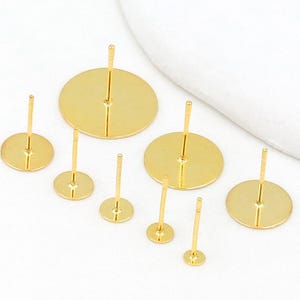 Peut inclure: Plusieurs clous d'oreilles dorés avec des pastilles circulaires de différentes tailles. Les tiges sont disposées sur une surface blanche. Les pastilles sont plates et conçues pour fixer des éléments décoratifs.