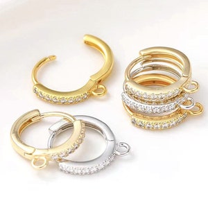 May include: Three pairs of gold and silver hoop earrings with a clear crystal embellishment. The earrings have a hinged closure and a small loop for attaching a charm.