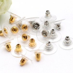 May include: A close-up of a group of earring backs. The backs are made of clear plastic with a metal post. Some of the posts are gold-colored and some are silver-colored.