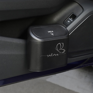 Puede incluir: Papelera negra para coche con un logotipo de mariposa y la palabra "salma" impresa en ella. La papelera tiene una tapa con botón pulsador y está montada en el lateral de una puerta de coche.