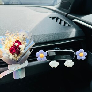 May include: A car air freshener with a bouquet of dried flowers and a purple flower design. The air freshener is attached to the car's air vent. The bouquet has a tag that says "Just for you".