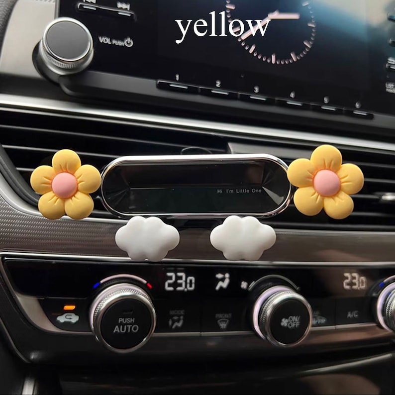 May include: A silver car air freshener with a yellow flower design and the text "Hi I'm Little One". The air freshener is attached to a car's air vent.