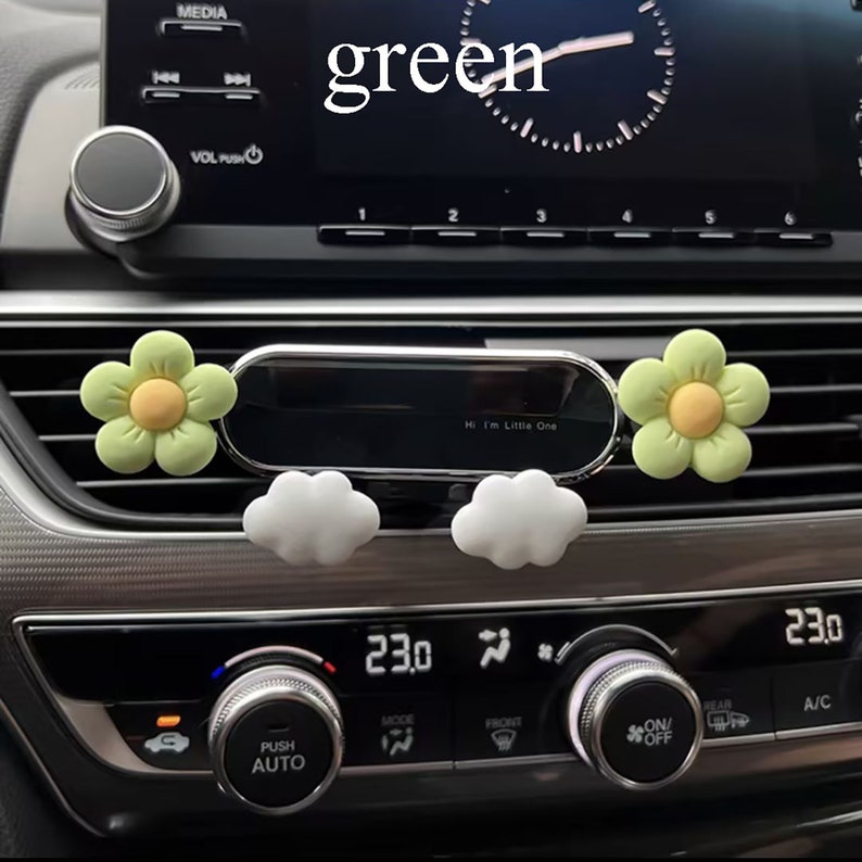 May include: A silver car phone holder with two green flower-shaped air fresheners and two white cloud-shaped air fresheners. The text "Hi I'm Little One" is visible on the phone holder.