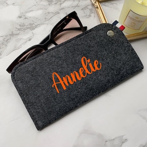 Puede incluir: Unas gafas de sol negras descansan sobre un estuche de fieltro gris oscuro. El estuche está personalizado con el nombre "Annelie" en naranja. Un pequeño broche plateado y un detalle de tela a rayas adornan la parte superior.