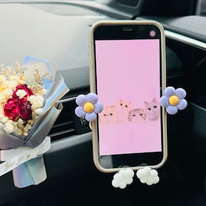 May include: A white phone holder with purple flower accents and white cloud-shaped bases. The phone holder is attached to a car vent and has a pink phone screen with a cartoon image of cats.