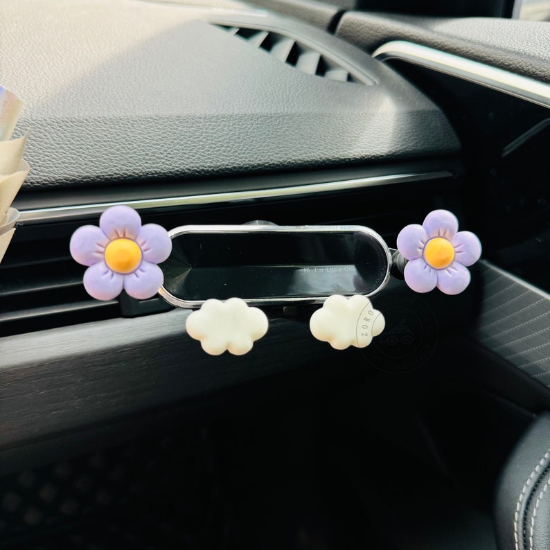 May include: A car air vent phone holder with a purple flower and white cloud design. The holder has a small mirror attached to it.