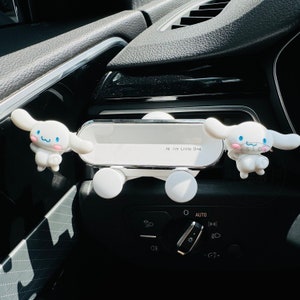 May include: White car phone holder with two white cartoon dog figures attached. The phone holder has the text "Hi I'm Little One" printed on it. The car holder is attached to the dashboard of a car.