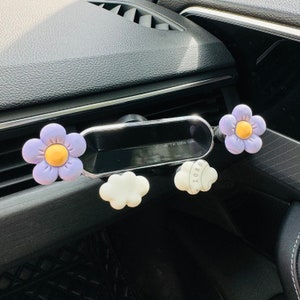May include: A car phone holder with a purple flower design. The holder has a small mirror and a cloud-shaped design. The text "Hi I'm Little Off" is visible on the mirror.