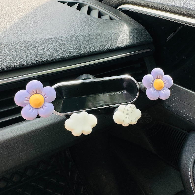 May include: A car phone holder with a purple flower design. The holder has a small mirror and a cloud-shaped design. The text "Hi I'm Little Off" is visible on the mirror.