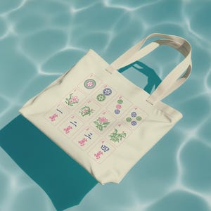 May include: Cream-colored tote bag featuring a Mahjong tile design. The design includes floral and geometric patterns in pink, green, and blue. The bag has cream-colored handles and is set against a blue water background.
