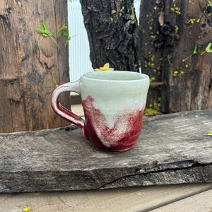 May include: A handmade ceramic mug with a white top and a deep red, abstract design on the bottom half. The handle is also red. The mug sits on a weathered wooden surface, suggesting a rustic aesthetic. The mug is ideal for coffee or tea.