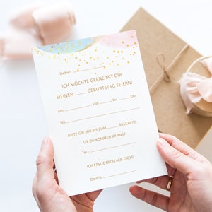 May include: A white fill-in-the-blank birthday invitation card with gold text and watercolor accents of blue and pink. The card is held by a person's hands, with a gift box in the background.