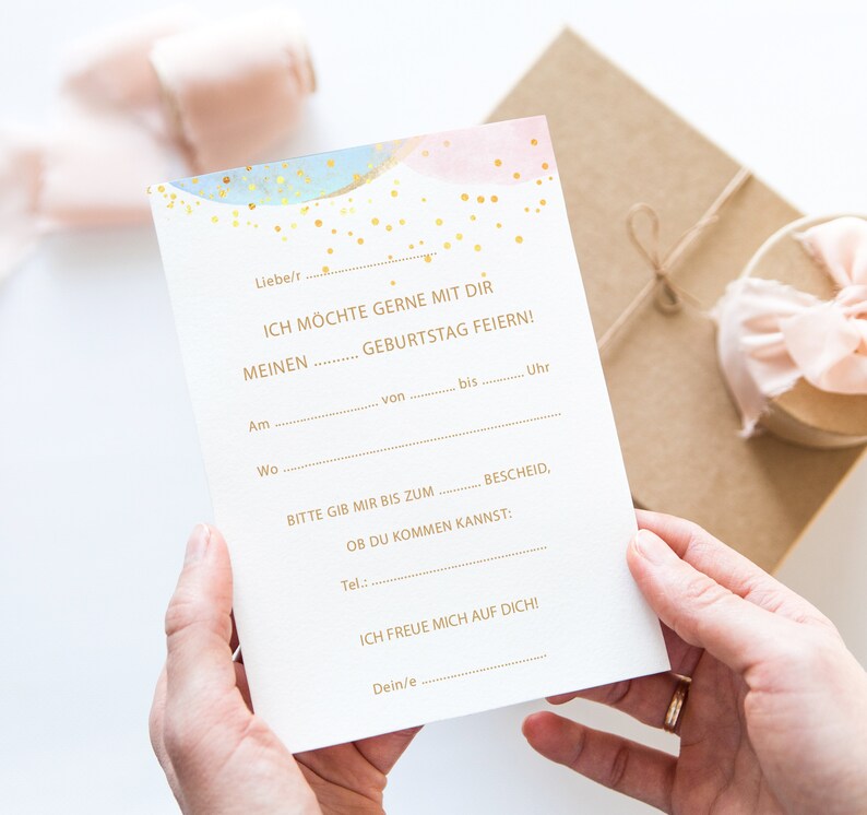 May include: A white fill-in-the-blank birthday invitation card with gold text and watercolor accents of blue and pink. The card is held by a person's hands, with a gift box in the background.