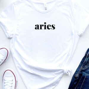 Op de afbeelding: Wit t-shirt met het woord "aries" in het zwart gedrukt. Het shirt wordt gecombineerd met witte sneakers met rode accenten en een blauwe jeans. Het t-shirt is in de taille geknoopt.