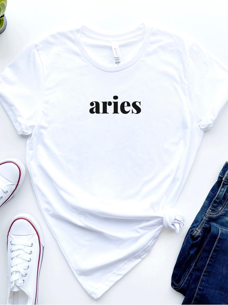 Op de afbeelding: Wit t-shirt met het woord "aries" in het zwart gedrukt. Het shirt wordt gecombineerd met witte sneakers met rode accenten en een blauwe jeans. Het t-shirt is in de taille geknoopt.