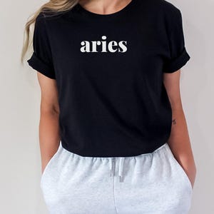 Op de afbeelding: Zwart T-shirt met het witte woord "aries" op de borst. Het shirt heeft korte mouwen en wordt gecombineerd met lichtgrijze joggingbroek. Het model draagt het shirt en de joggingbroek.