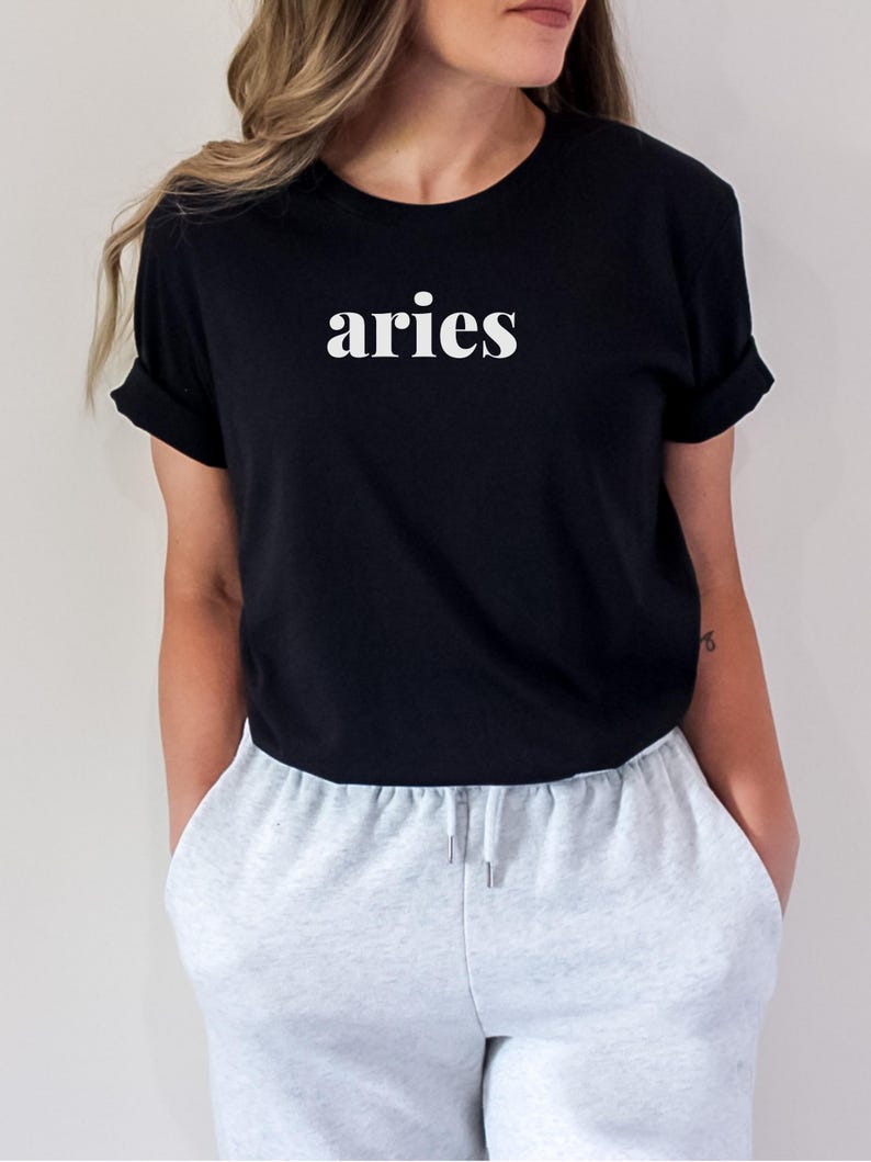 Op de afbeelding: Zwart T-shirt met het witte woord "aries" op de borst. Het shirt heeft korte mouwen en wordt gecombineerd met lichtgrijze joggingbroek. Het model draagt het shirt en de joggingbroek.