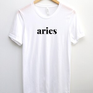 Op de afbeelding: Een wit t-shirt met het woord "aries" in zwarte letters, hangend aan een houten hanger. Het shirt heeft een ronde hals en korte mouwen en is gemaakt van een zachte, lichte stof.
