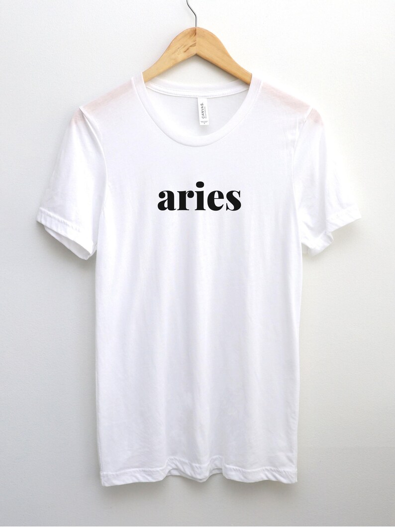 Op de afbeelding: Een wit t-shirt met het woord "aries" in zwarte letters, hangend aan een houten hanger. Het shirt heeft een ronde hals en korte mouwen en is gemaakt van een zachte, lichte stof.