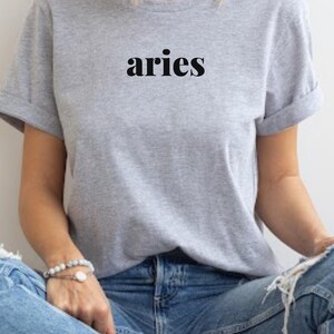 Op de afbeelding: Een lichtgrijs t-shirt met het woord "aries" in zwarte letters. De mouwen zijn opgerold. Het t-shirt wordt gedragen met een blauwe jeans. Een armband is zichtbaar op de pols.