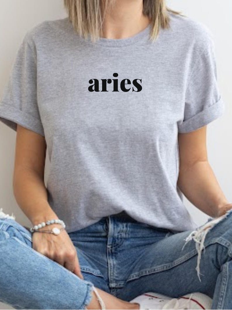 Op de afbeelding: Een lichtgrijs t-shirt met het woord "aries" in zwarte letters. De mouwen zijn opgerold. Het t-shirt wordt gedragen met een blauwe jeans. Een armband is zichtbaar op de pols.