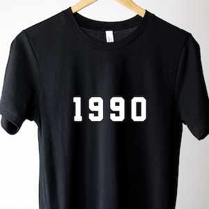 Puede incluir: Camiseta negra con texto blanco que dice "1990" en una fuente en negrita y en bloque.