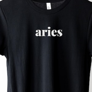 Op de afbeelding: Zwart t-shirt met het woord "aries" in het wit gedrukt, gecentreerd op de borst. Het shirt hangt aan een houten hanger. De tekst is in een eenvoudige, schreefloze lettertype. De achtergrond is wit.