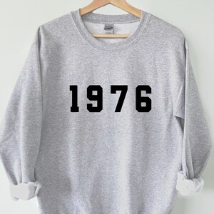 Könnte beinhalten: Ein hellgraues Sweatshirt mit Rundhalsausschnitt und dem schwarzen Schriftzug 1976, der in einer fetten Blockschrift mittig auf der Brust aufgedruckt ist. Das Kleidungsstück hat lange Ärmel mit gerippten Bündchen und einen gerippten Halsausschnitt.