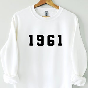 May include: White sweatshirt featuring the year "1961" in bold black lettering. The garment is suspended from a wooden hanger. The Gildan brand label is visible at the neckline.