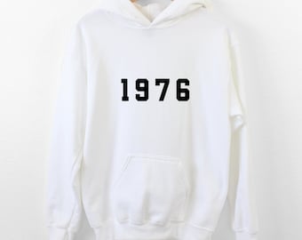 Könnte beinhalten: Ein weißer Kapuzenpullover mit langen Ärmeln und dem schwarzen Text 1976, der in einer fetten Blockschrift mittig auf der Brust aufgedruckt ist. Das Kleidungsstück verfügt über eine Kängurutasche, eine Kapuze mit Kordelzug sowie Rippbündchen.