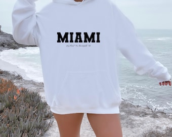 Może przedstawiać: Biała bluza z kapturem z nadrukiem "MIAMI" w czarnych, pogrubionych literach i współrzędnymi "25.7617° N, 80.1918° W" nadrukowanymi poniżej.