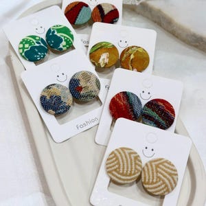 May include: Five pairs of circular fabric-covered button earrings displayed on white cards labeled Fashion. The earrings feature diverse patterns including green floral, mustard yellow, blue and tan abstract, red and teal stripes, and beige chevron.