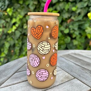 May include: A clear glass tumbler filled with iced coffee, featuring a bamboo lid and a bright pink silicone straw. The exterior is decorated with colorful printed patterns of heart-shaped and round concha sweet bread in orange, pink, and cream.