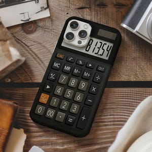 May include: Black phone case designed to look like a calculator, with a screen displaying the number 81354. The case has calculator buttons and a camera lens cutout. The case is on a wooden surface.
