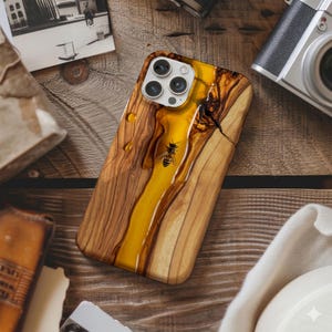 May include: A wooden phone case with a yellow resin river design and a bee embedded in the resin. The case has a natural wood grain pattern and a glossy finish. The case is designed for a smartphone.