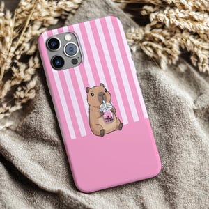 May include: Pink phone case with a vertical striped pattern and a solid pink bottom. The case features a cartoon capybara drinking a pink bubble tea. The phone case is designed to fit a smartphone.