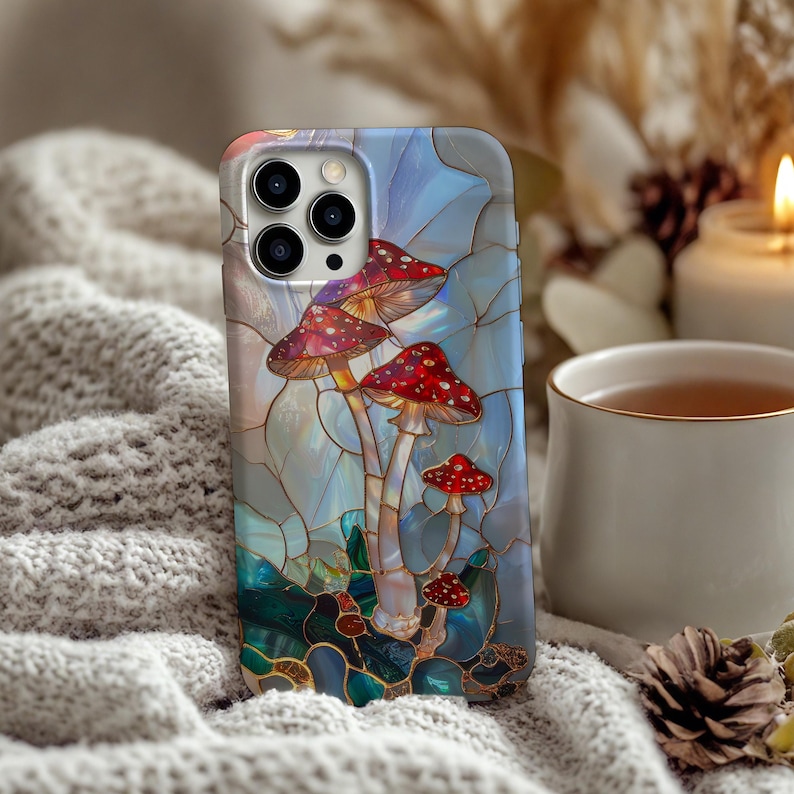 K&ouml;nnte beinhalten: Eine Handyh&uuml;lle mit einem Buntglas-Design, das rot-wei&szlig; gepunktete Pilze zeigt. Die H&uuml;lle hat einen blau-gr&uuml;nen Hintergrund mit Perleffekt. Die Handyh&uuml;lle liegt auf einer weichen, wei&szlig;en, gestrickten Oberfl&auml;che.