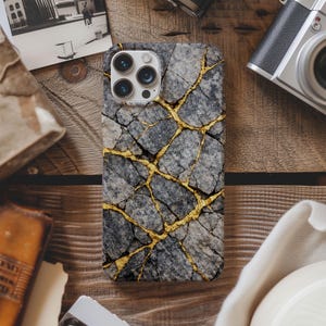May include: A smartphone case with a gray stone pattern and gold-colored vein-like accents. The case has a multi-lens camera cutout. The case is on a wooden surface with other objects.