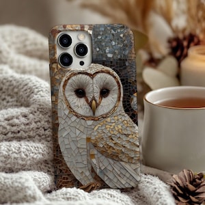 May include: A mosaic-style phone case featuring a white owl with gold accents. The owl is centered on a background of brown and gray mosaic tiles. The case is resting on a soft, white, textured surface.