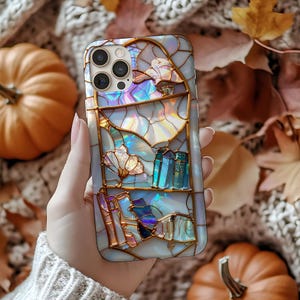 Peut inclure: Coque de t&eacute;l&eacute;phone nacr&eacute;e avec un motif de vitrail. Le bo&icirc;tier pr&eacute;sente un paysage avec des livres, des montagnes et des fleurs dans des tons de bleu, d'or et de couleurs iris&eacute;es. Le bo&icirc;tier est tenu dans une main.