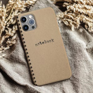 May include: A tan phone case featuring a spiral notebook design. The word "notebook" is printed in black. The phone's camera lenses are visible at the top. The case rests on a grey fabric.