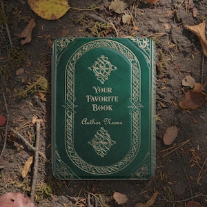 May include: A hardcover book with a dark green cover and gold embossed detailing. The text "YOUR FAVORITE BOOK" and "Author Name" are displayed on the cover. The book is lying on a bed of leaves and soil.